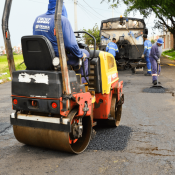 A imagem mostra trabalhadores da EMDEF (Empresa Municipal para o Desenvolvimento de Franca) realizando recapeamento asfáltico em uma rua. Um operário opera um rolo compactador enquanto outros espalham o asfalto com pás. Ao fundo, uma máquina distribui o material. Todos usam uniformes azuis com identificação da EMDEF.