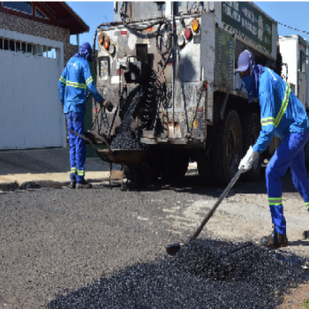 Dois funcionários da EMDEF com uniformes azuis, um está operando o caminhão com o asfalto e o outro alinhando o asfalto na rua.